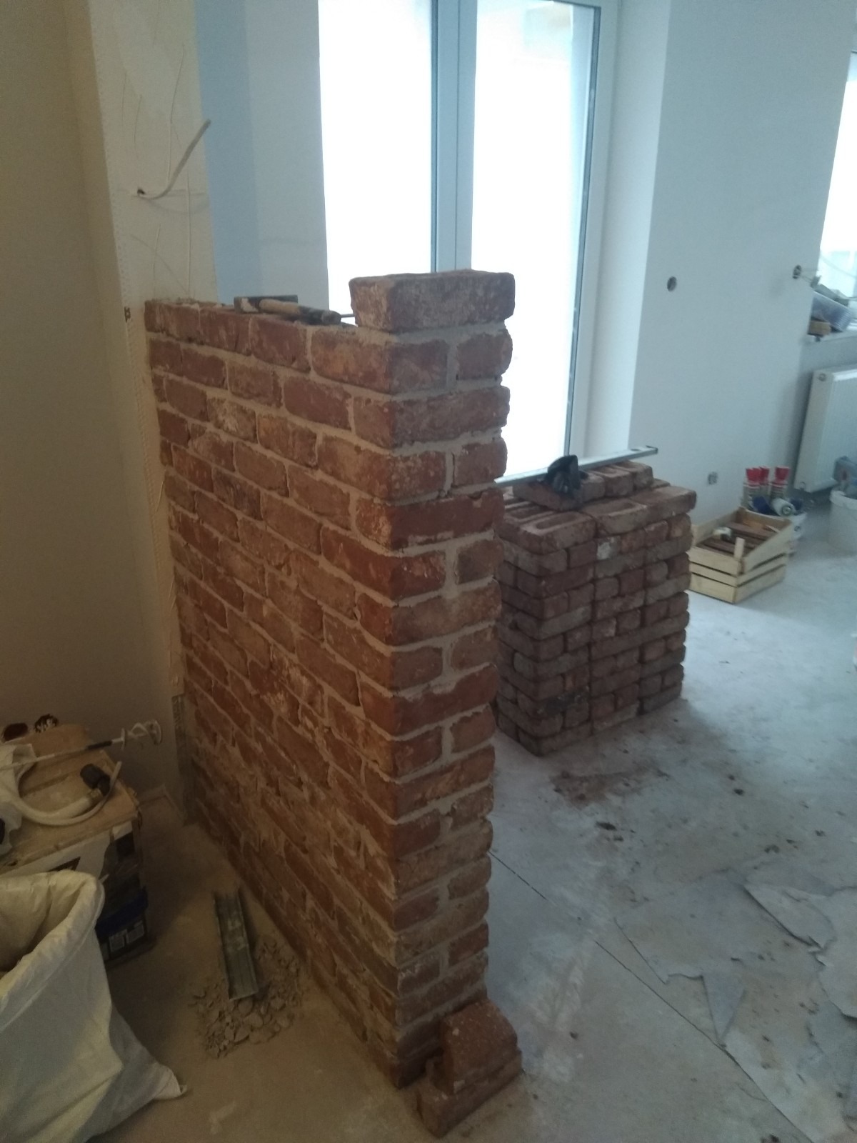 Wznoszenie ścianki działowej z czerwonej cegły w surowym wnętrzu, widoczny stos cegieł i narzędzia, etap budowy przed wykończeniem.