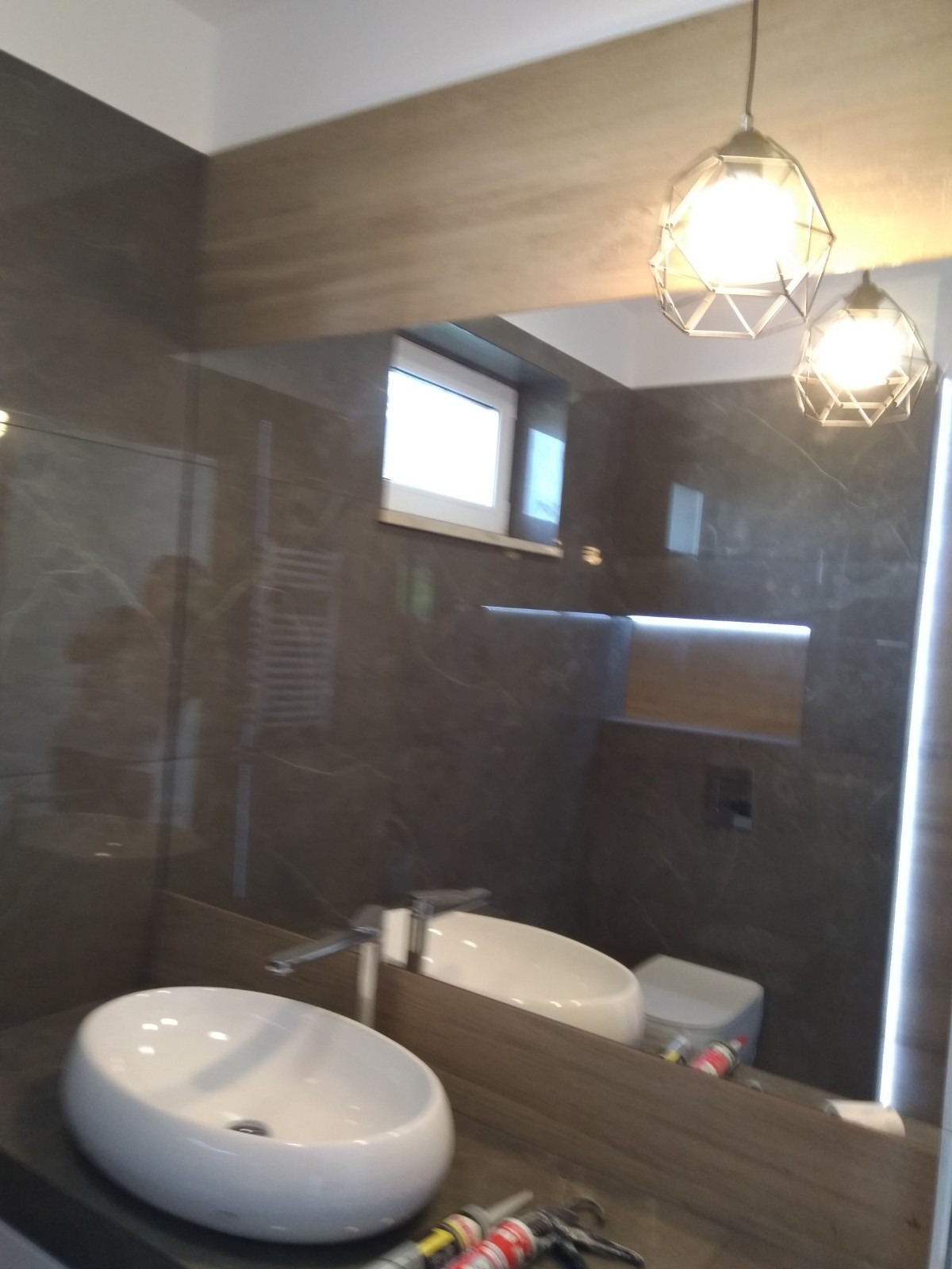 Elegancka łazienka z ciemnymi płytkami imitującymi marmur, owalna umywalka nablatowa, bateria chromowana, oświetlenie punktowe w geometrycznych oprawach, widoczne narzędzia i silikon w tle...