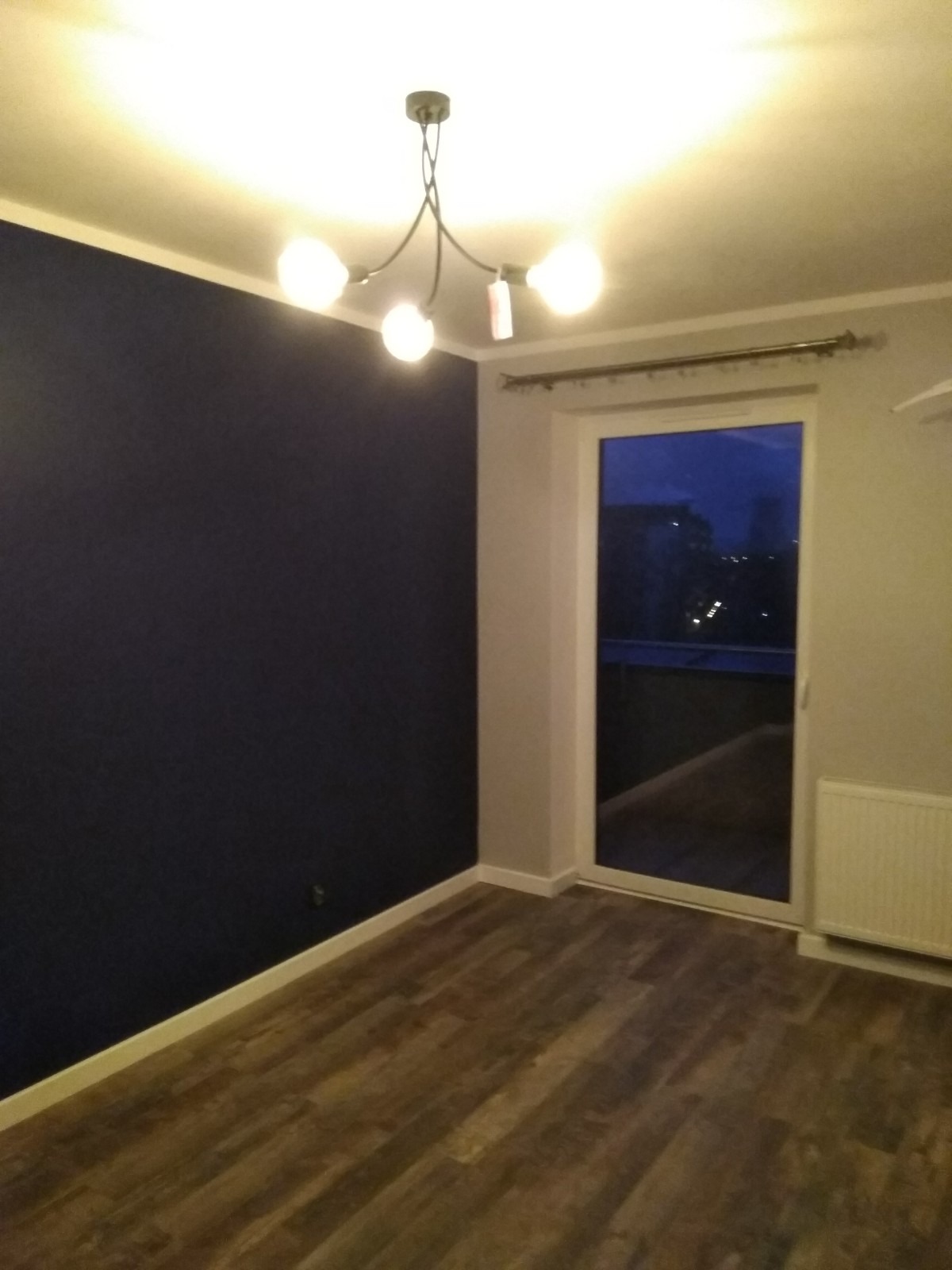 Wnętrze pokoju po remoncie: granatowa ściana, drewnopodobne panele podłogowe, biała listwa przypodłogowa, nowoczesna lampa sufitowa z kulistymi żarówkami, okno balkonowe z widokiem na nocne miasto.