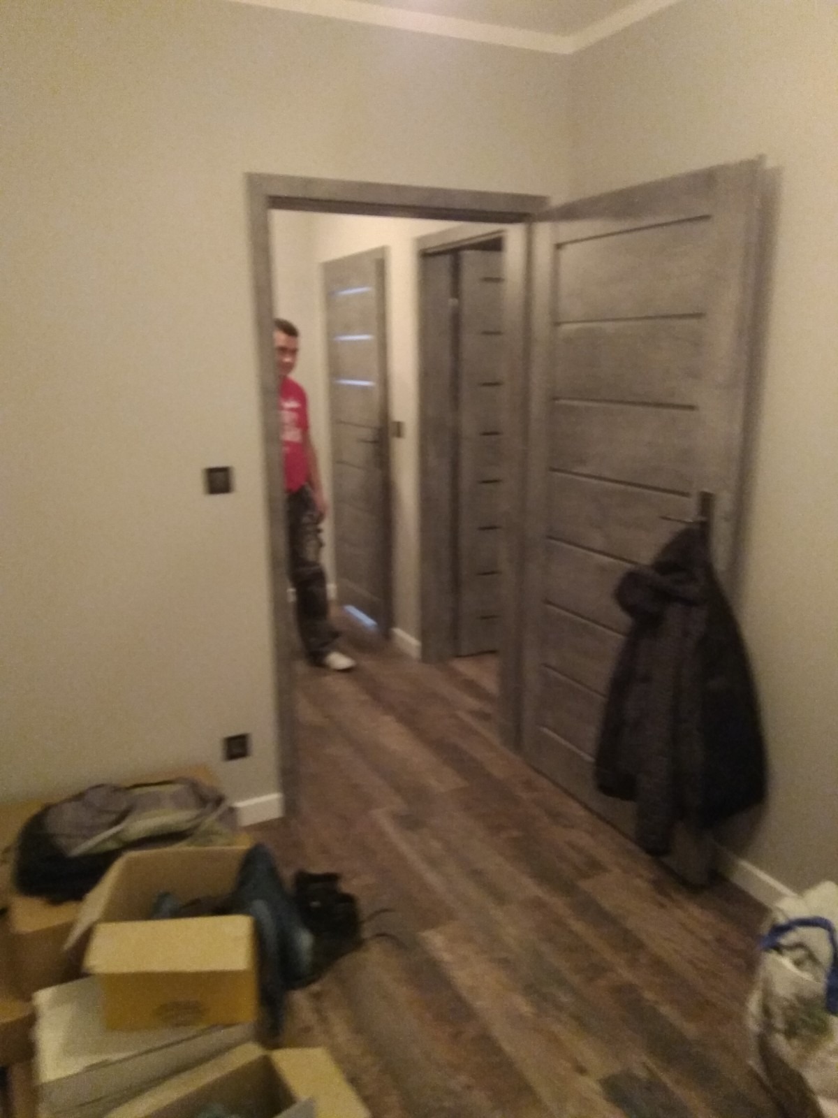 Montaż szarych drzwi wewnętrznych z poziomymi frezowaniami, widoczny fragment korytarza z panelami podłogowymi, w tle majster w czerwonej koszulce.