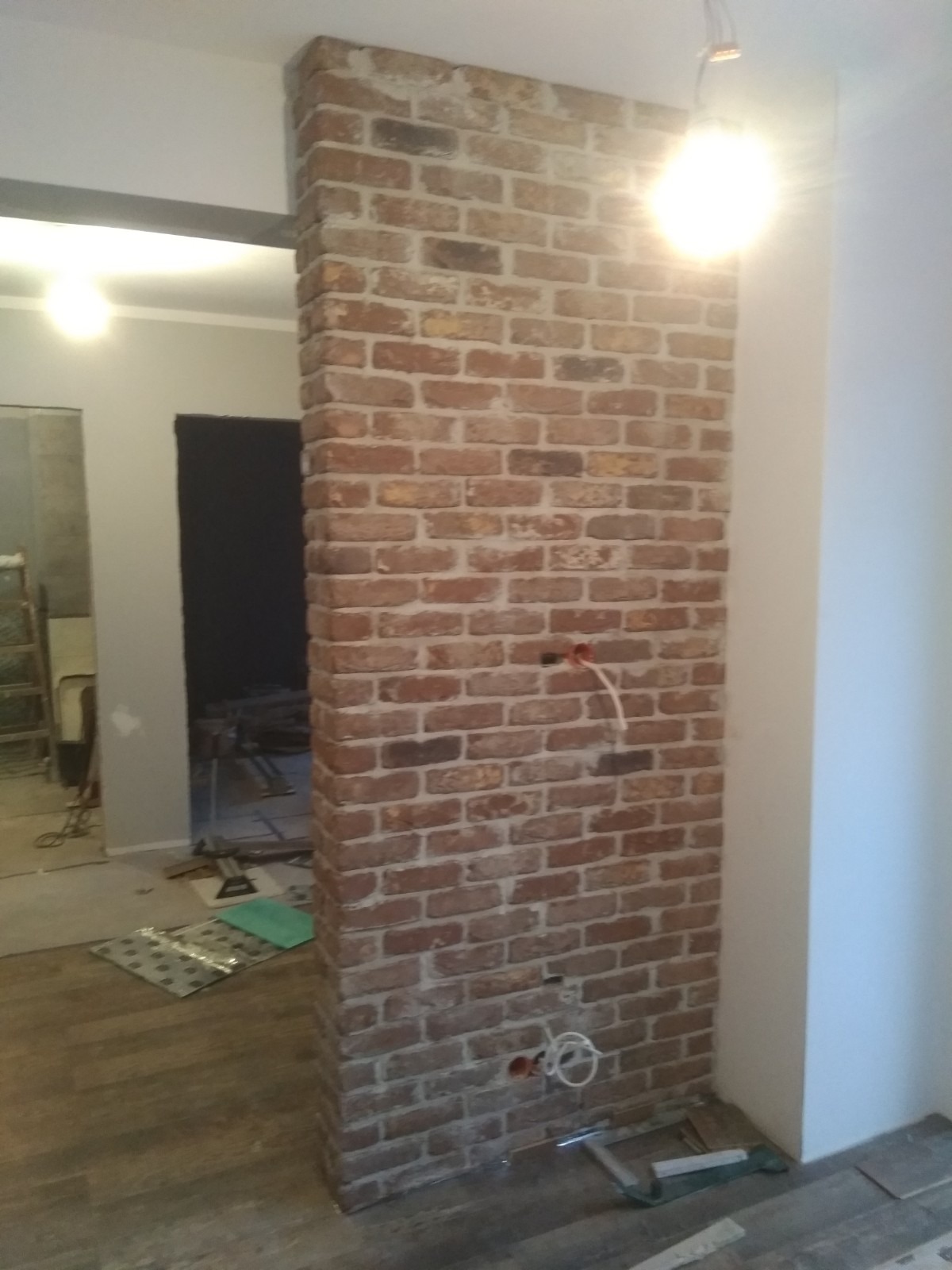 Wzniesiona ściana z cegły klinkierowej w trakcie prac wykończeniowych; widoczne wystające kable elektryczne, fragment podłogi z paneli i materiały budowlane w tle.