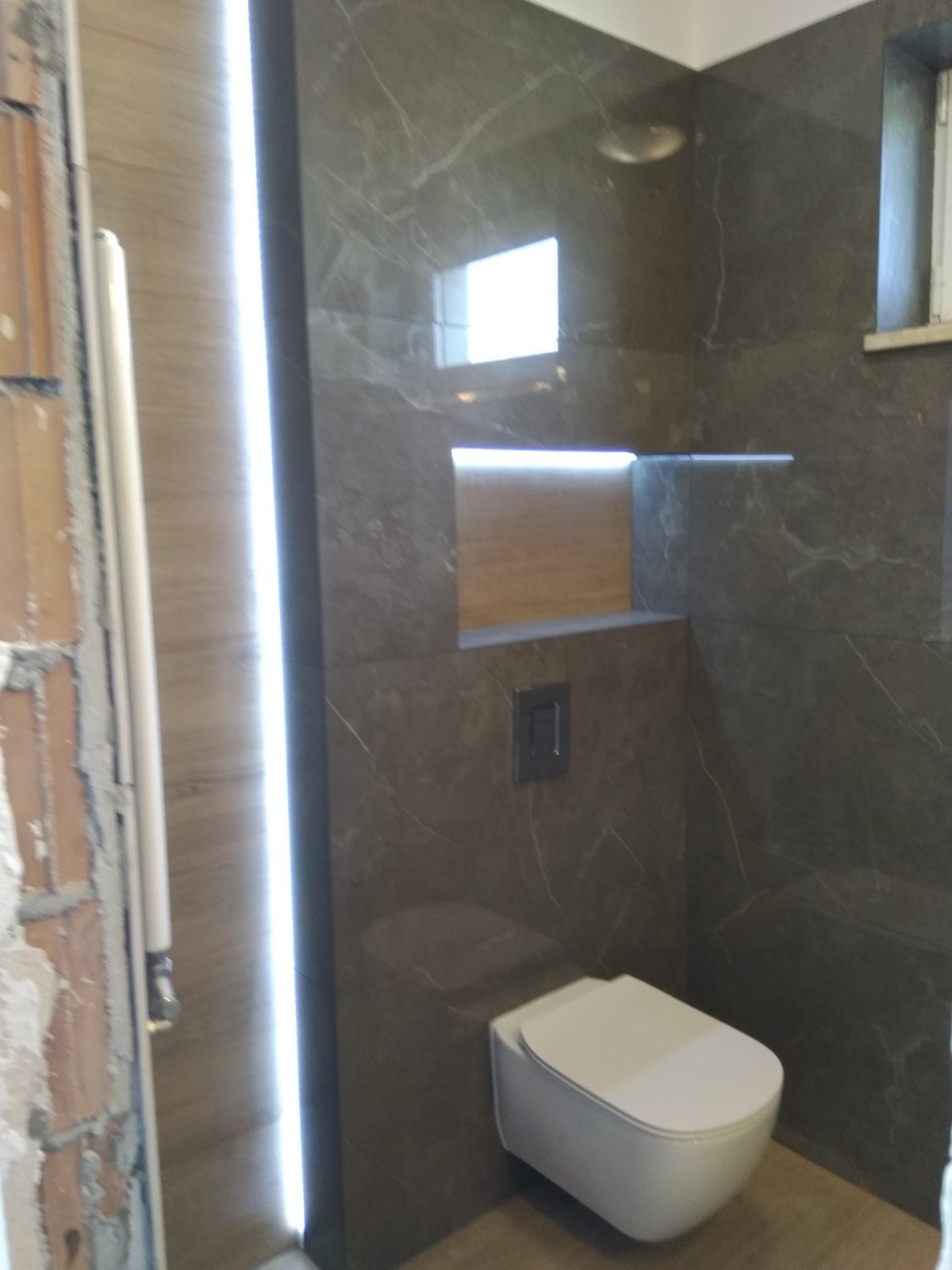Nowoczesna łazienka z ciemnymi płytkami imitującymi marmur, podwieszana toaleta, oświetlona wnęka i pionowe oświetlenie LED przy ścianie z surowej cegły po lewej stronie.