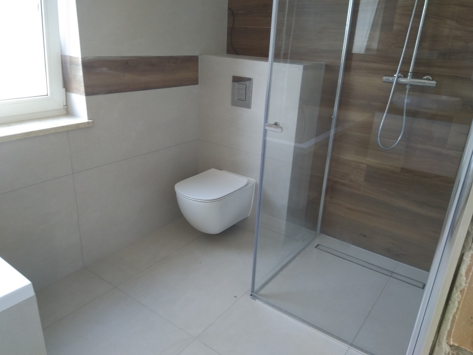 Nowoczesna łazienka z podwieszaną miską WC, szklaną kabiną prysznicową typu walk-in i drewnopodobnymi akcentami na ścianach.