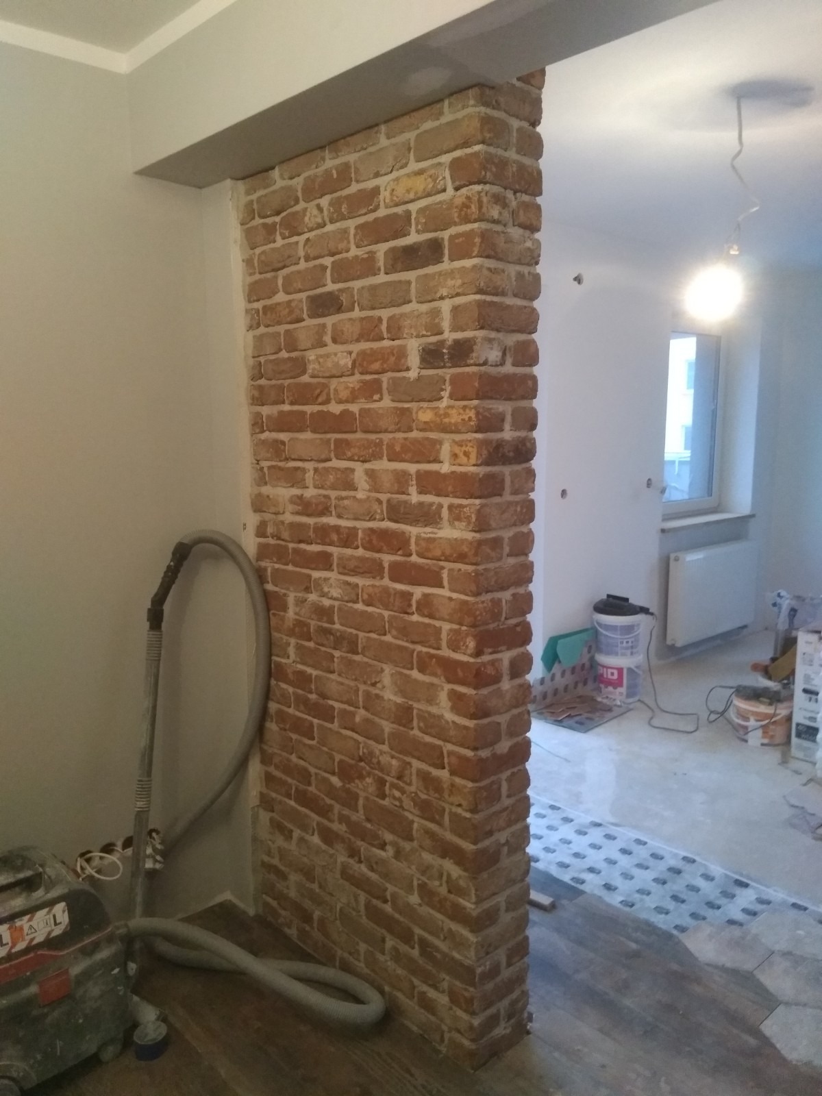 Ceglana ścianka działowa w trakcie prac wykończeniowych; widoczny odkurzacz przemysłowy, podłoga częściowo wyłożona panelami, ściany przygotowane do malowania.