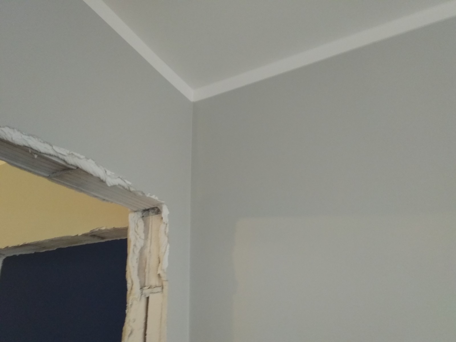 Fragment ściany pomalowanej na szaro z białym paskiem dekoracyjnym pod sufitem i surowym ościeżem drzwi w trakcie prac wykończeniowych, widoczne ślady zaprawy.