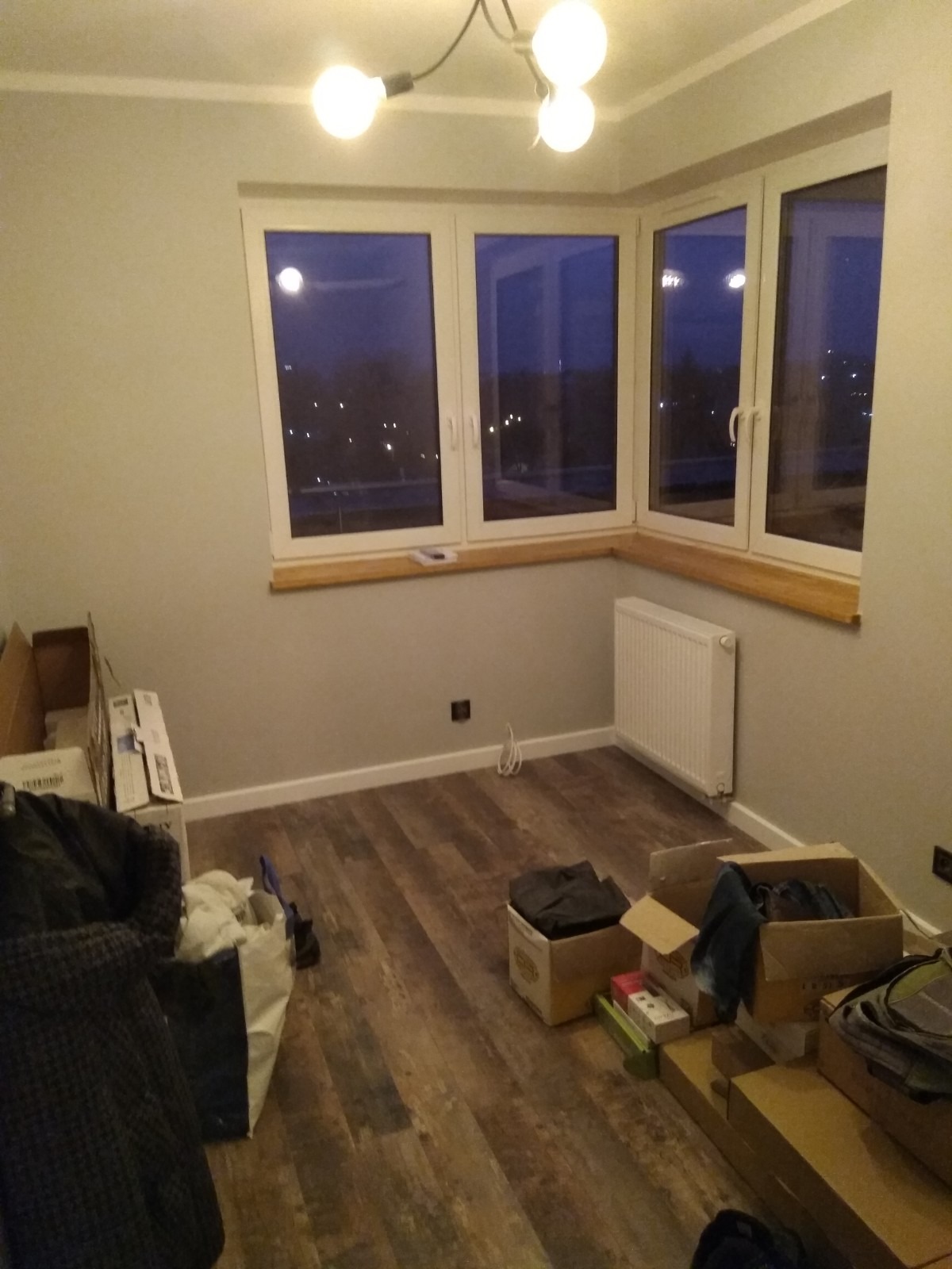 Pokój po remoncie z nową podłogą z paneli imitujących drewno, grzejnikiem, trzema oknami z widokiem na nocne miasto i surową żarówką na suficie, częściowo zagracony kartonami.