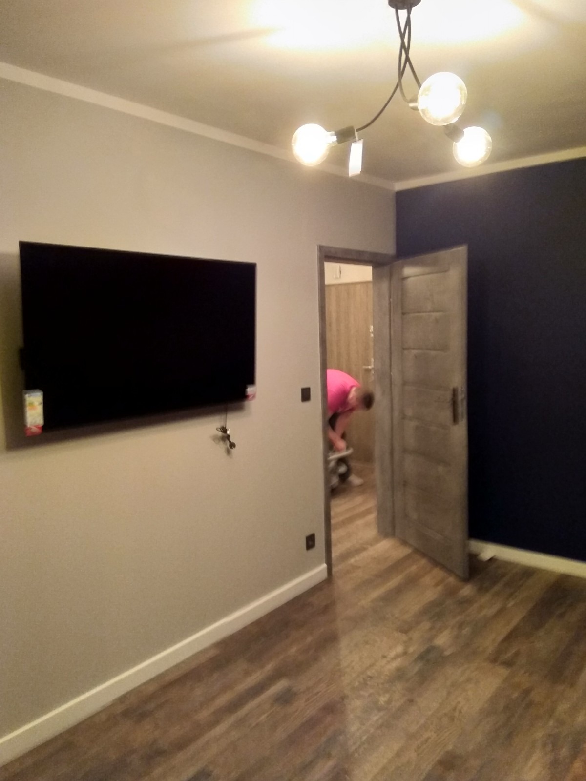 Pokój po remoncie: drewnopodobne panele podłogowe, szara ściana z zawieszonym telewizorem, fragment ściany pomalowany na granatowo, widoczna osoba w różowej koszulce pracująca w oddali, nowoczesna...