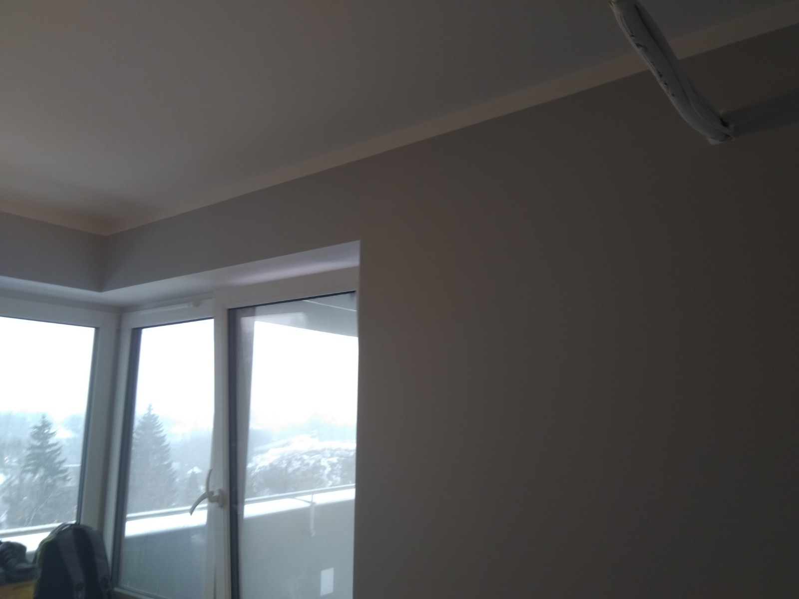 Świeżo pomalowana ściana w odcieniach szarości z widocznym oknem balkonowym i fragmentem sufitu, z wystającym przewodem instalacji elektrycznej w rogu.