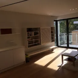 Jasne wnętrze salonu z parkietem, minimalistyczną zabudową meblową, przeszklonym wyjściem na balkon i wpadającym przez okno światłem słonecznym.