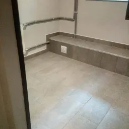 Wykafelkowane pomieszczenie z szarymi płytkami podłogowymi i niewielkim oknem, widoczne szare rury przy ścianie, wentylacja w dolnej części ściany.