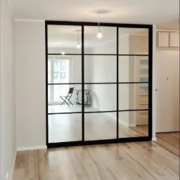 Przeszklona ściana działowa z czarnymi szprosami, oddzielająca przestrzeń mieszkalną, widok na minimalistyczny pokój z krzesłem i stolikiem.