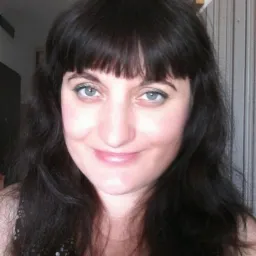 Portret uśmiechniętej kobiety z ciemnymi włosami i niebieskimi oczami, z delikatnym makijażem i grzywką, w bluzce w białe kropki.