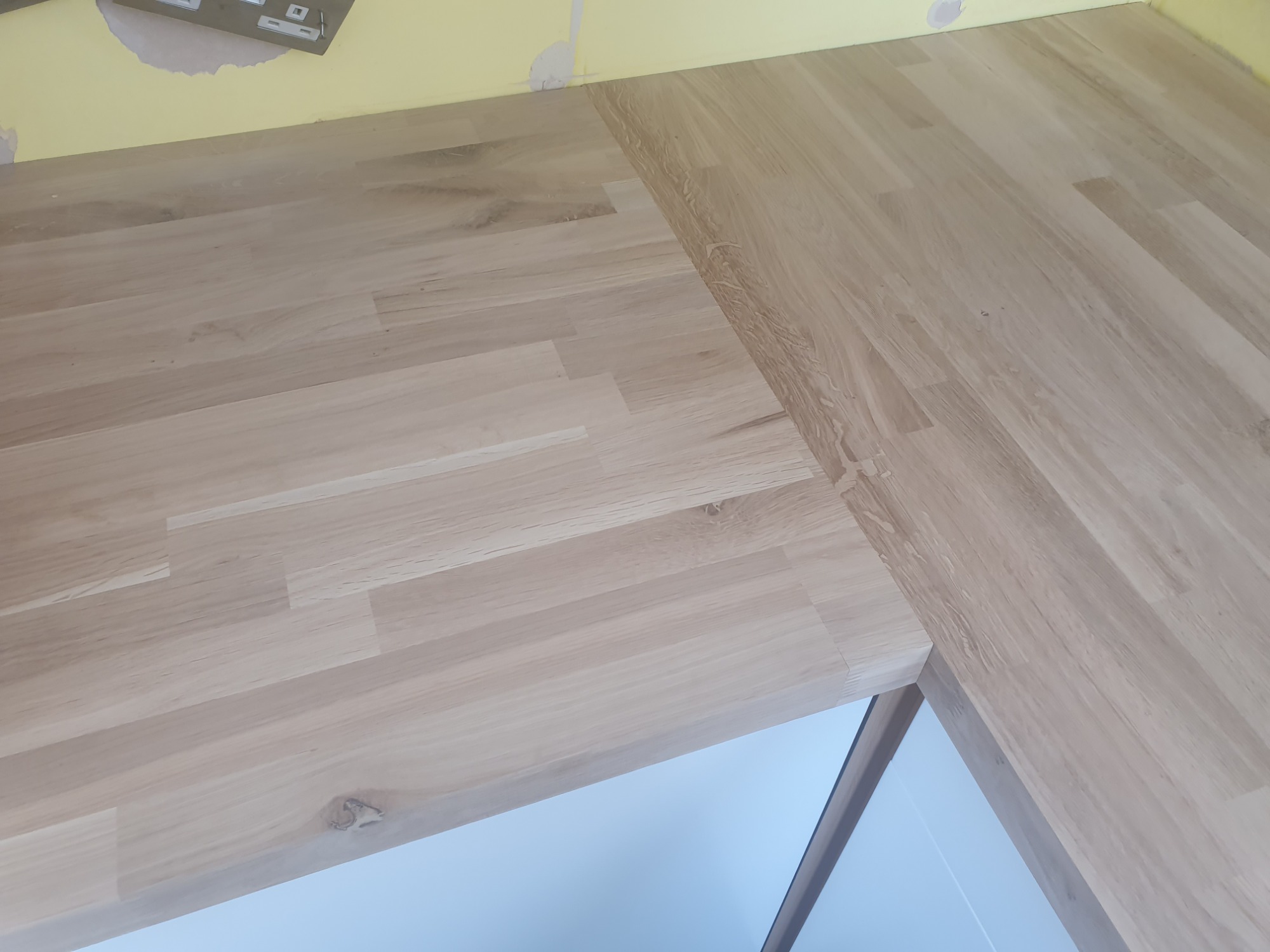 Fragment blatu z jasnego drewna klejonego, widoczny na tle żółtej ściany z niedokończonymi pracami malarskimi i białej szafki.