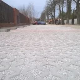 Szara, ażurowa nawierzchnia z kostki betonowej wypełniona drobnym kruszywem, prowadząca wzdłuż ogrodzenia z cegły klinkierowej i drewnianych sztachet, z widocznymi samochodami i budynkiem w tle.