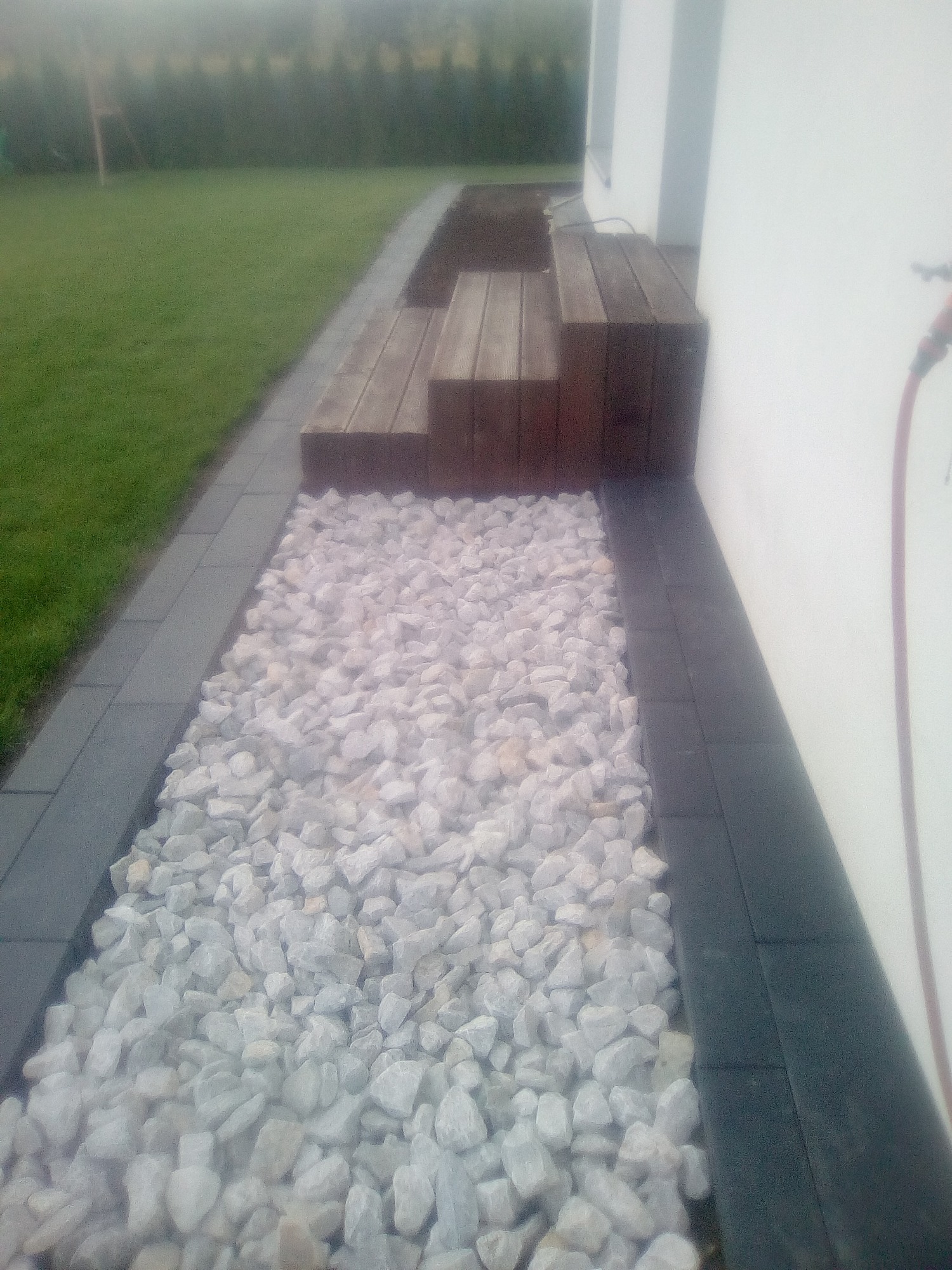 Schody z drewnianych belek i wysypany żwir ozdobny obok ściany domu w Łodzi, obrzeże z kostki brukowej.