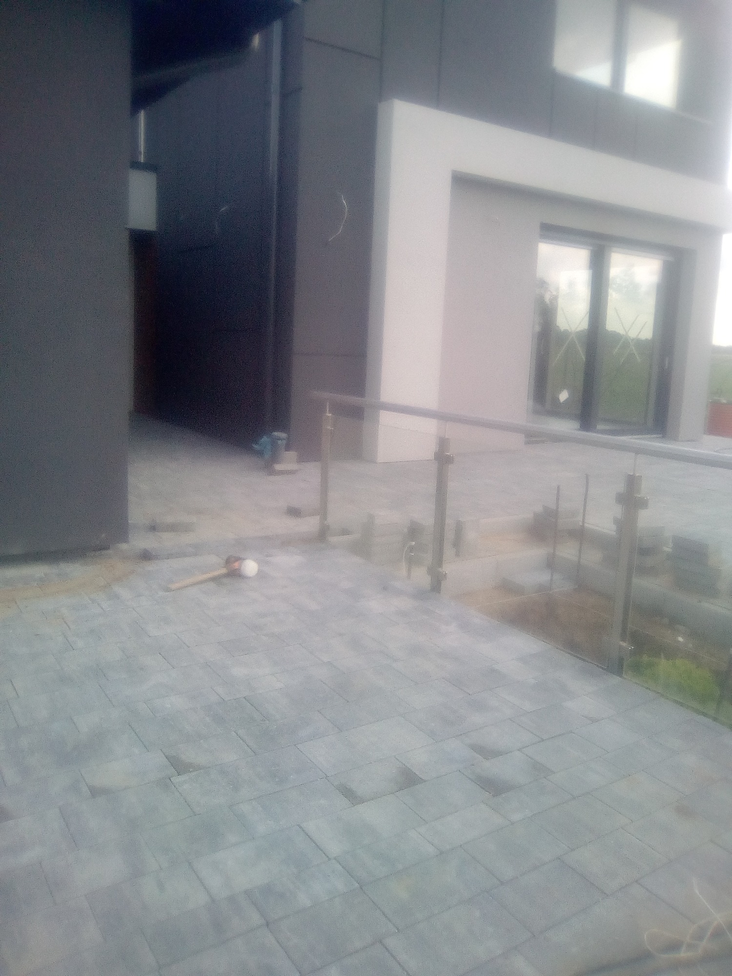 Nowoczesny taras z szarej kostki brukowej, częściowo wykończony, z szklaną balustradą i widocznym młotkiem brukarskim.