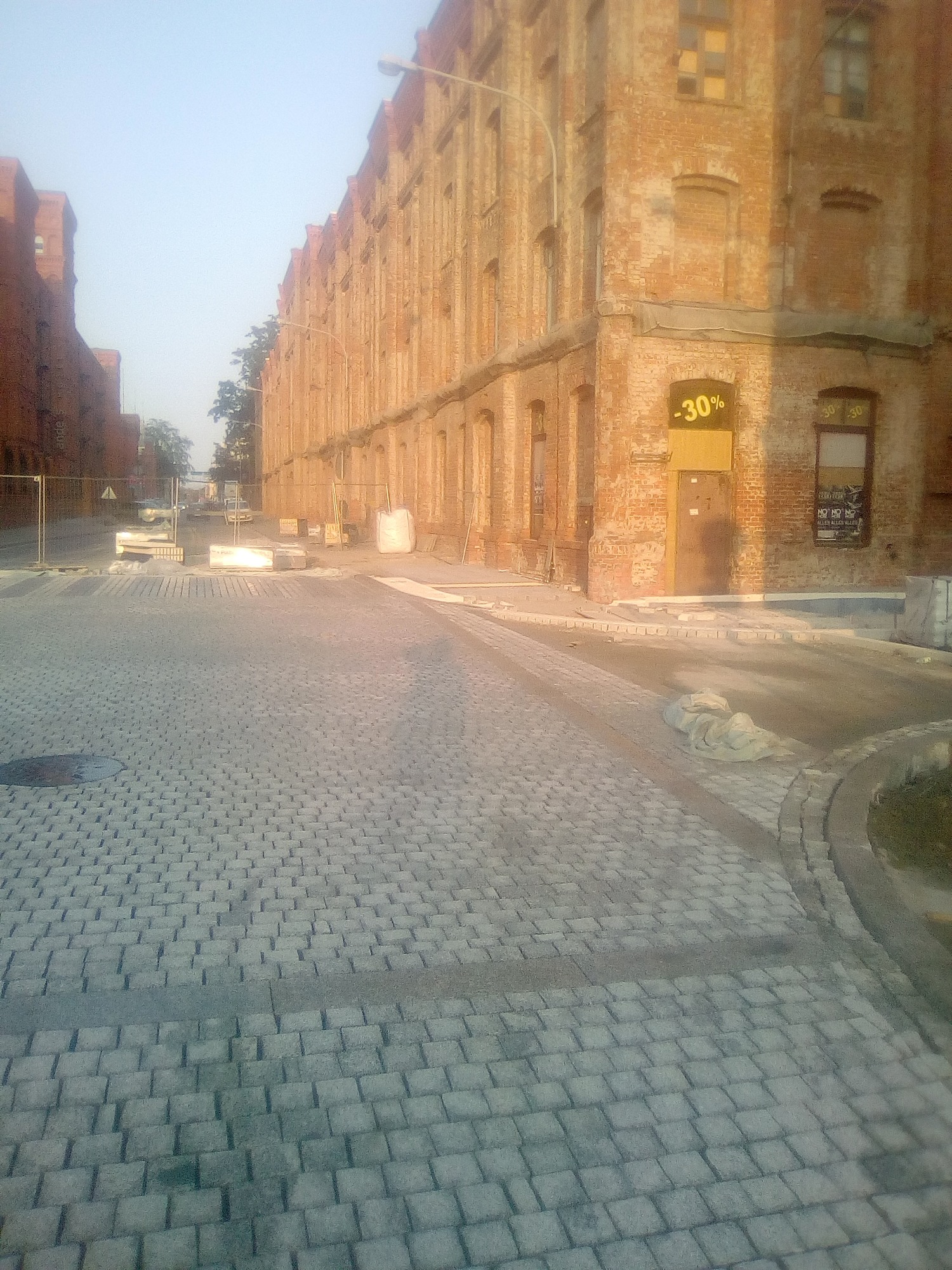 Ulica wyłożona szarą kostką brukową, w tle ceglane budynki z żółtym znakiem -30% na jednym z nich, widoczne zabezpieczenia robót drogowych.