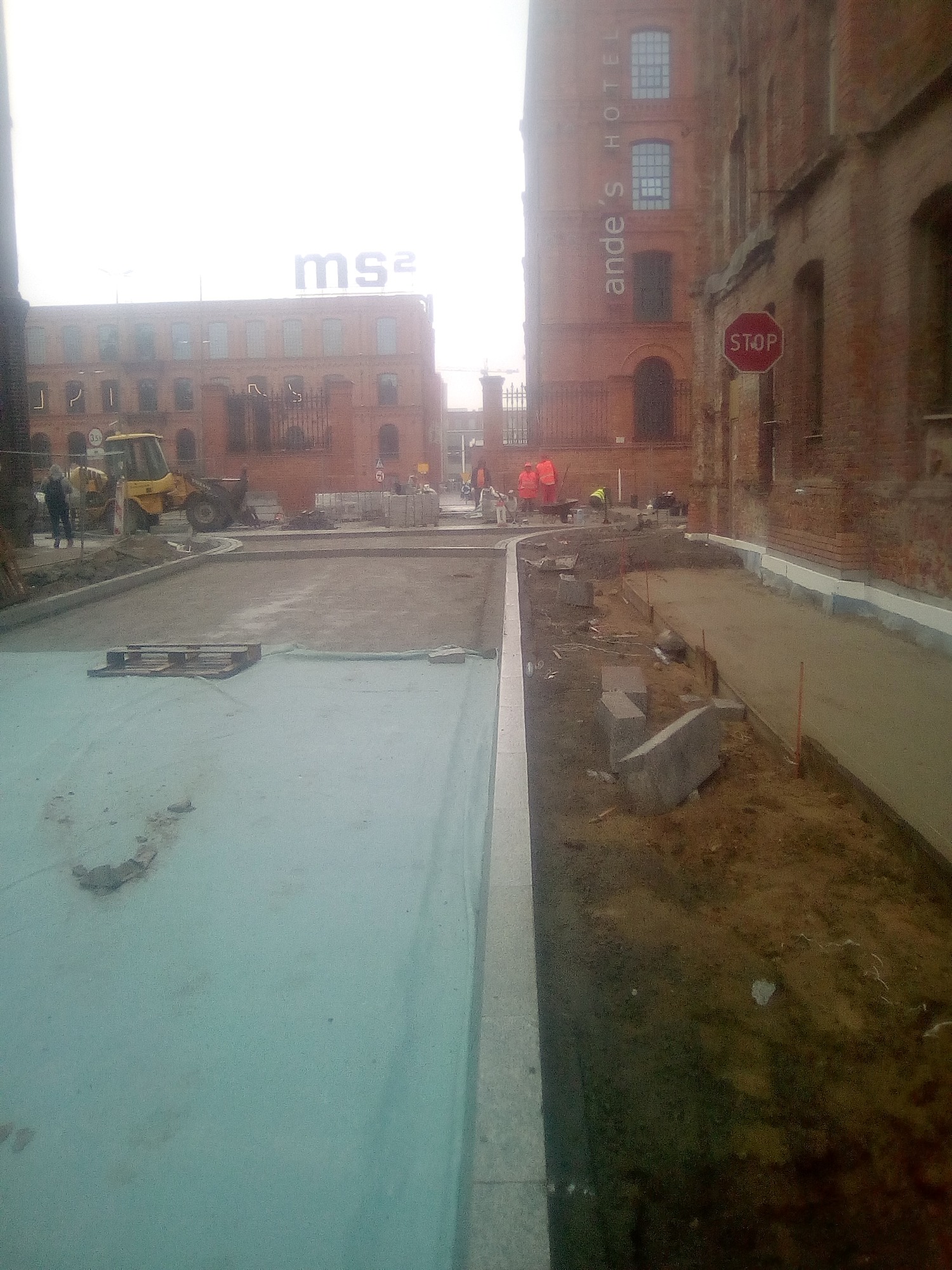 Plac budowy z żółtą koparką w tle, robotnicy w pomarańczowych kamizelkach układają obrzeża chodnika, w tle budynki z czerwonej cegły.