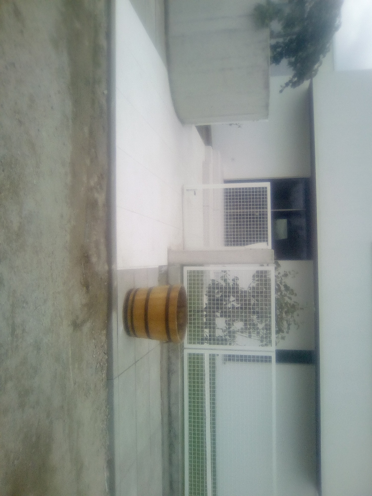 Drewniana beczka dekoracyjna na jasnoszarej kostce brukowej, białe ogrodzenie z kratką w tle, ujęcie z boku.