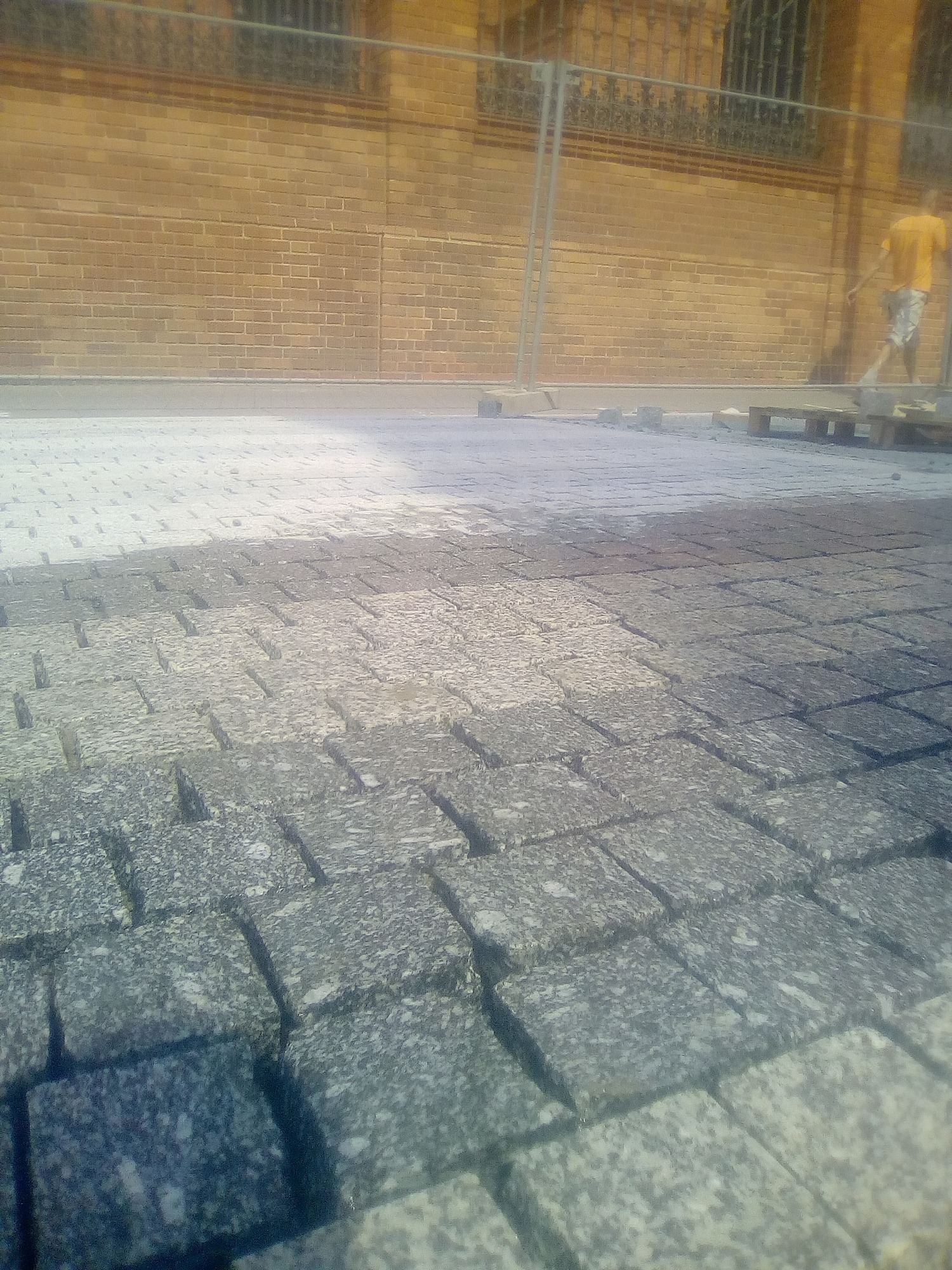 Fragment układanej nawierzchni z kostki granitowej w odcieniach szarości, z widocznym fragmentem muru z cegły w tle oraz pracownikiem w oddali.