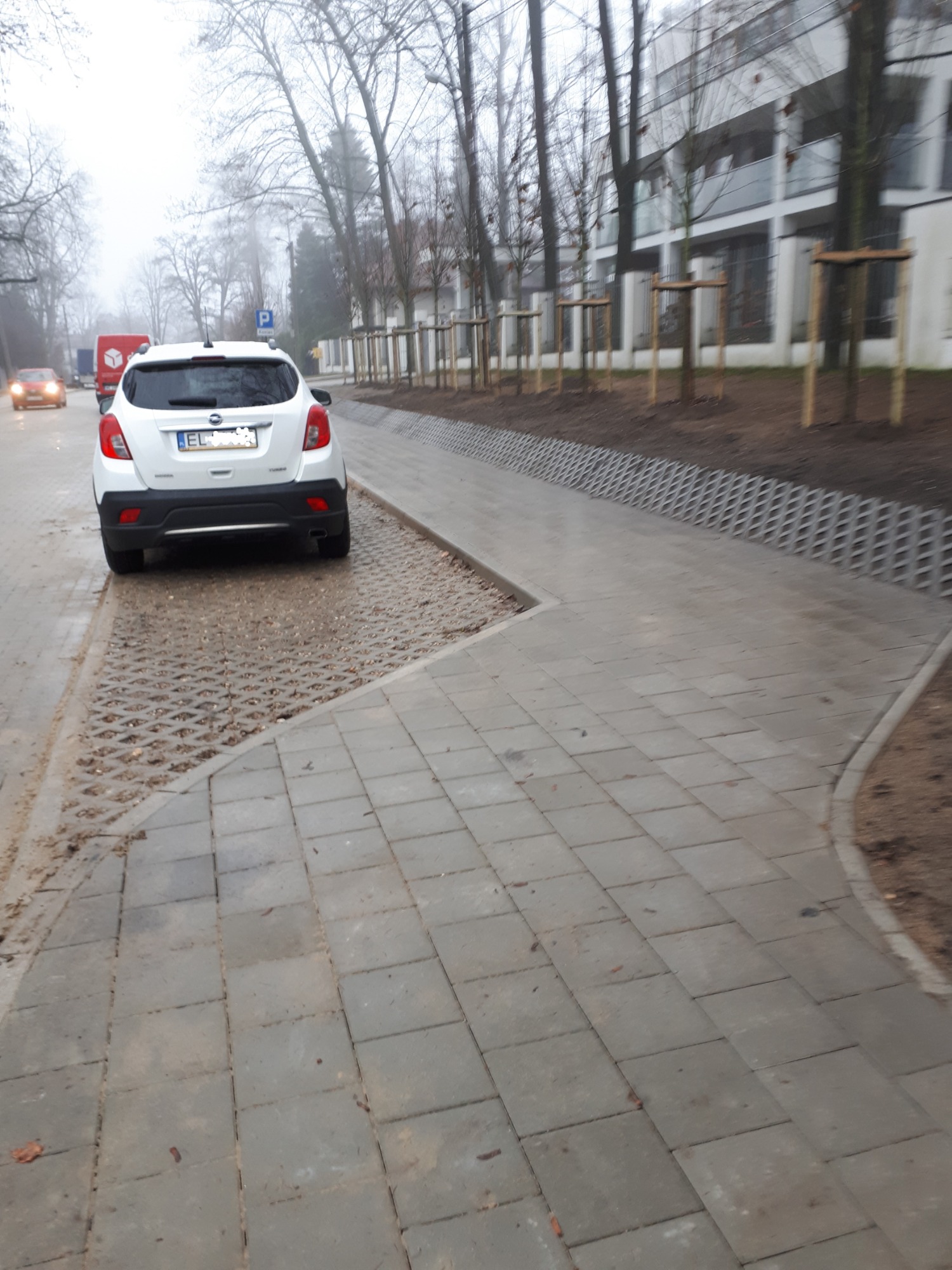 Szara kostka brukowa ułożona na ścieżce, z fragmentem ażurowej nawierzchni, obok zaparkowany biały samochód w pochmurny dzień.