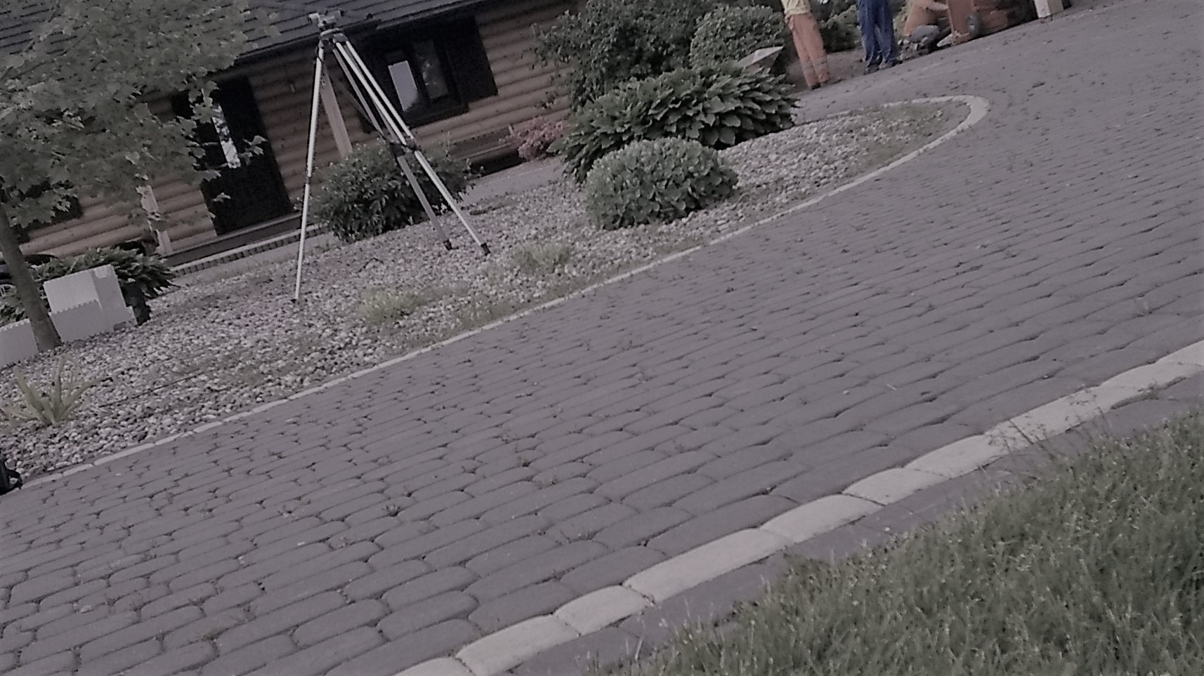Ułożona szara kostka brukowa prowadząca do drewnianego domu, widoczny trójnóg fotograficzny i fragment pracujących osób w tle.