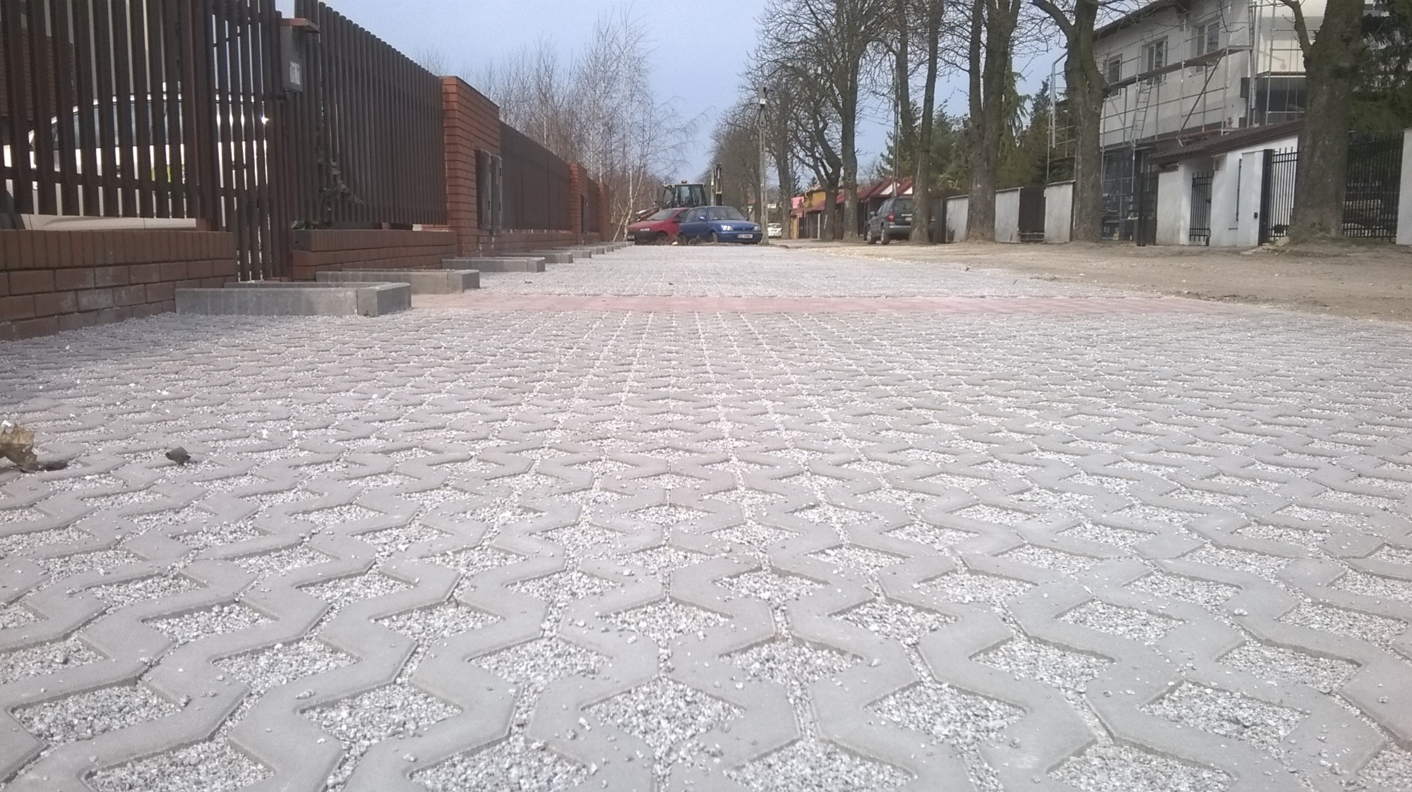 Szara, ażurowa nawierzchnia z kostki betonowej wypełniona drobnym kruszywem, prowadząca wzdłuż ogrodzenia z cegły klinkierowej i drewnianych sztachet, z widocznymi samochodami i budynkiem w tle.