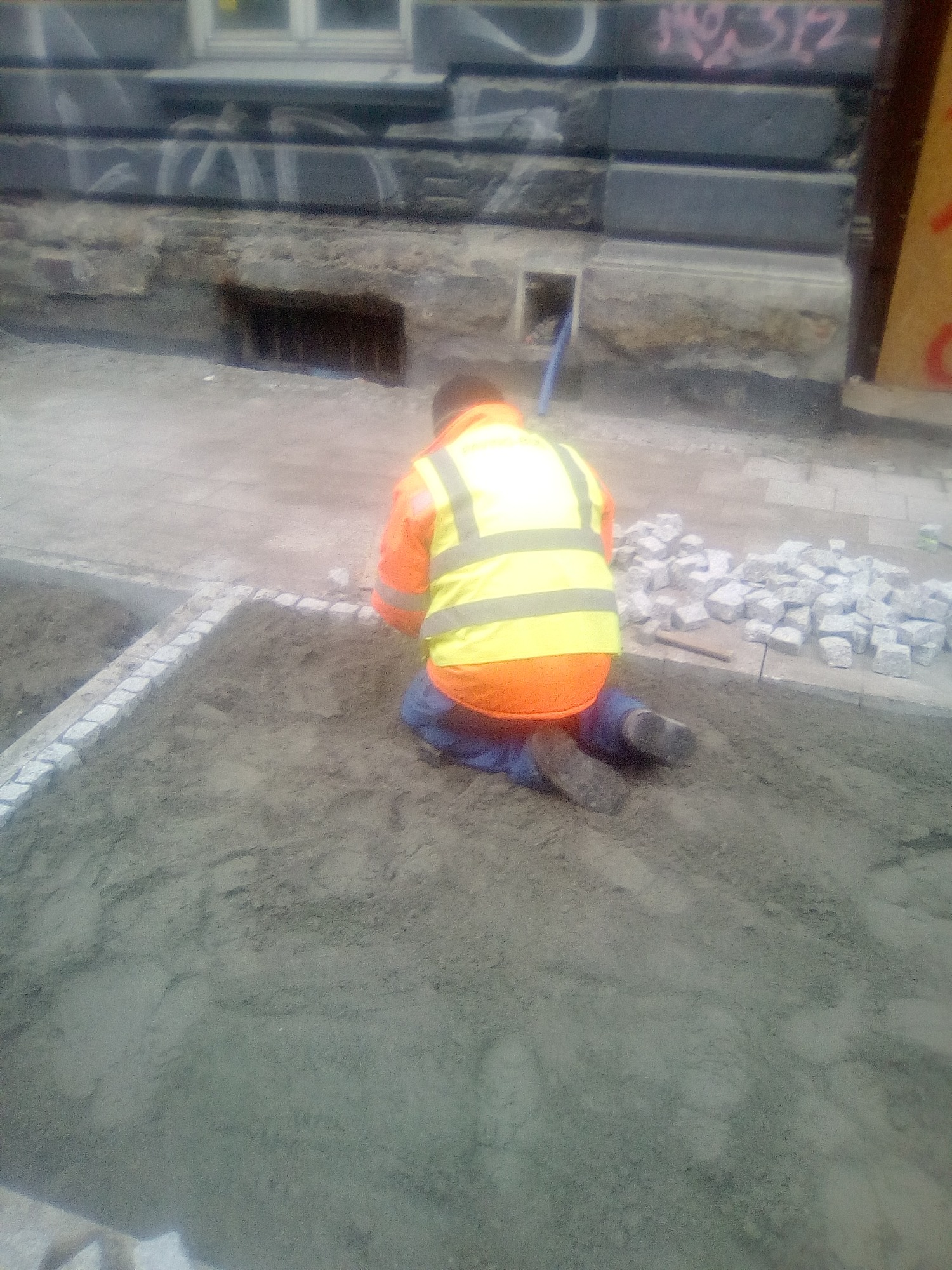 Pracownik w odblaskowej kamizelce klęczy układając obrzeże z kostki granitowej w przygotowanym podłożu na tle budynku z graffiti.