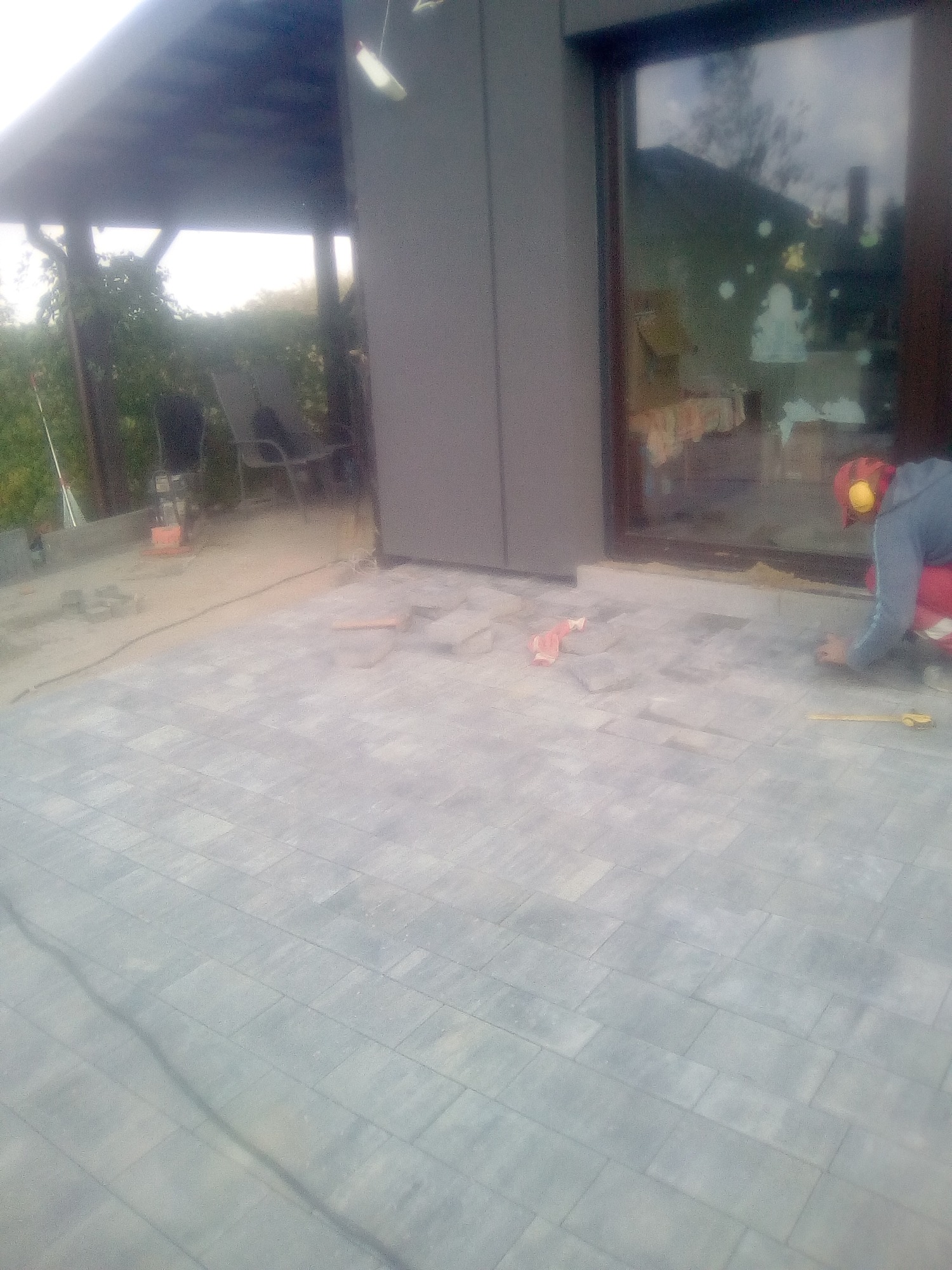 Prace brukarskie w toku: układanie jasnoszarej kostki brukowej na tarasie przed domem, pracownik w ubraniu roboczym z żółtymi słuchawkami ochronnymi przykuca obok okna, widoczny zarys altany w tle.