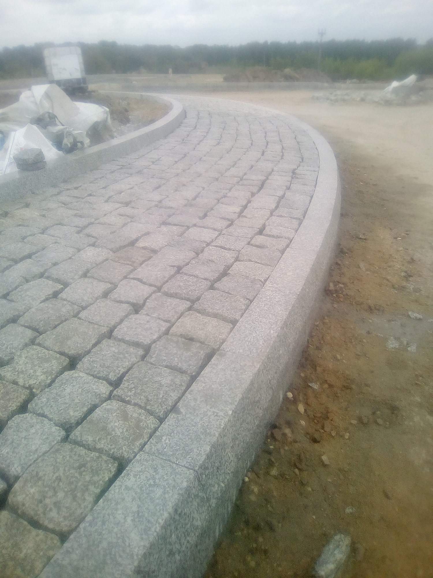 Nowo ułożona, zakrzywiona ścieżka z kostki granitowej z szarą obwódką, widok z boku na plac budowy w pochmurny dzień.