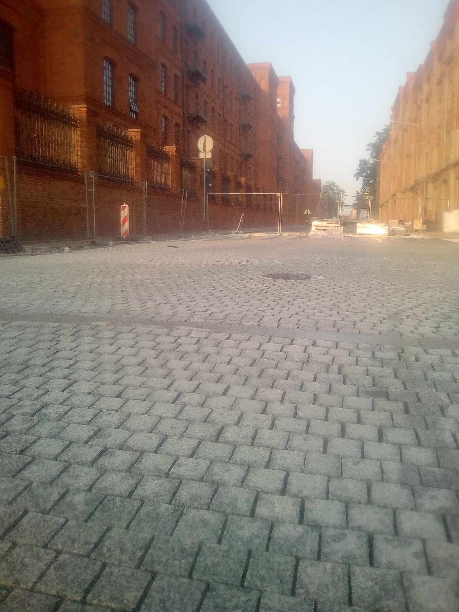 Ulica wyłożona szarą kostką brukową w otoczeniu ceglanych budynków z metalowymi ogrodzeniami i znakami drogowymi.