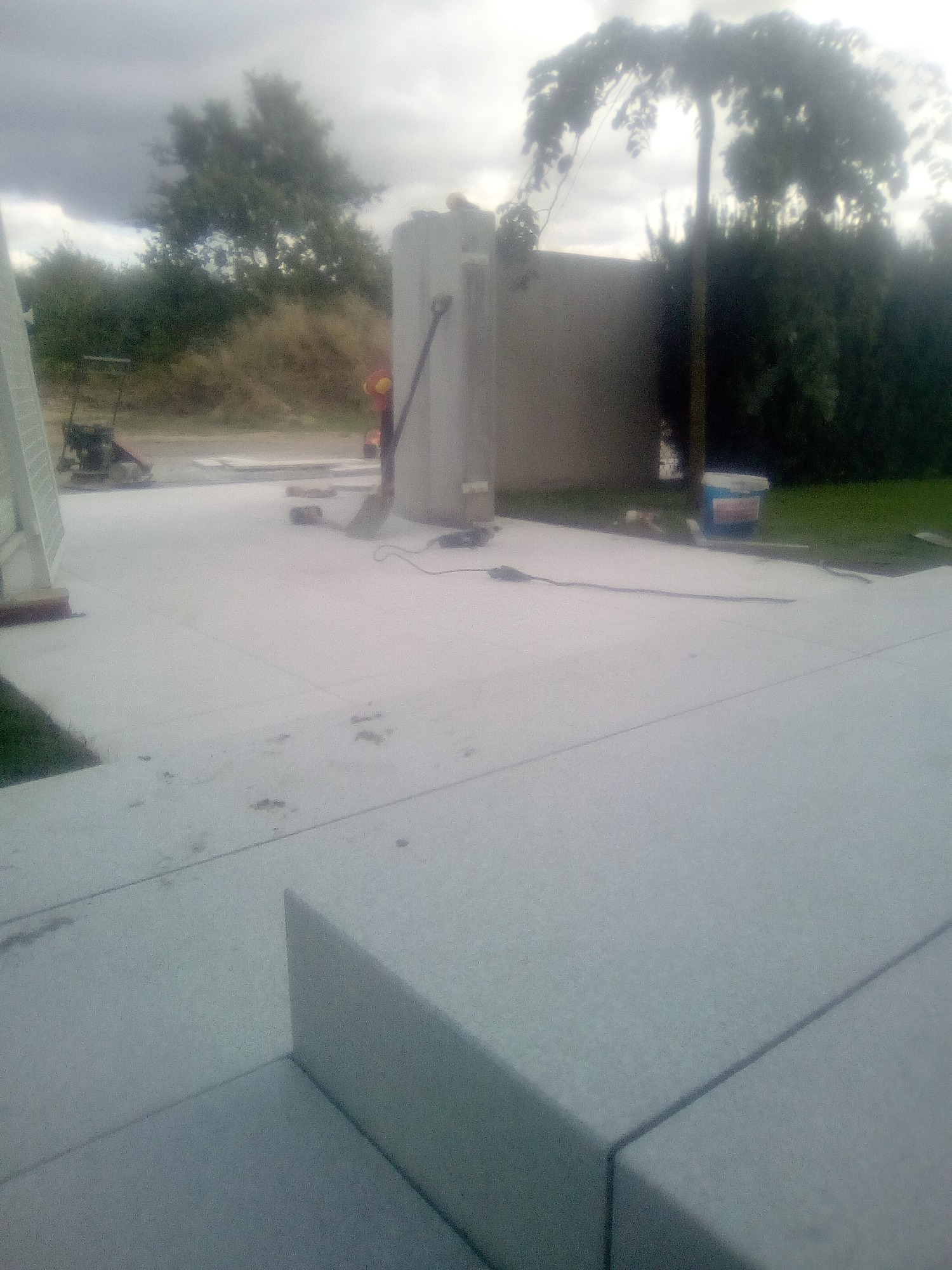 Szara, betonowa powierzchnia tarasu w trakcie budowy, widoczne narzędzia i materiały budowlane, w tle zieleń i pochmurne niebo.