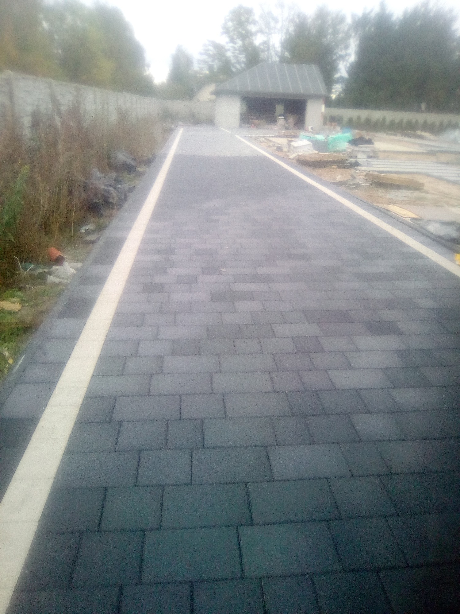 Nowo ułożona, długa ścieżka z ciemnoszarej kostki brukowej z jasnymi obrzeżami, prowadząca do budynku w budowie w tle.