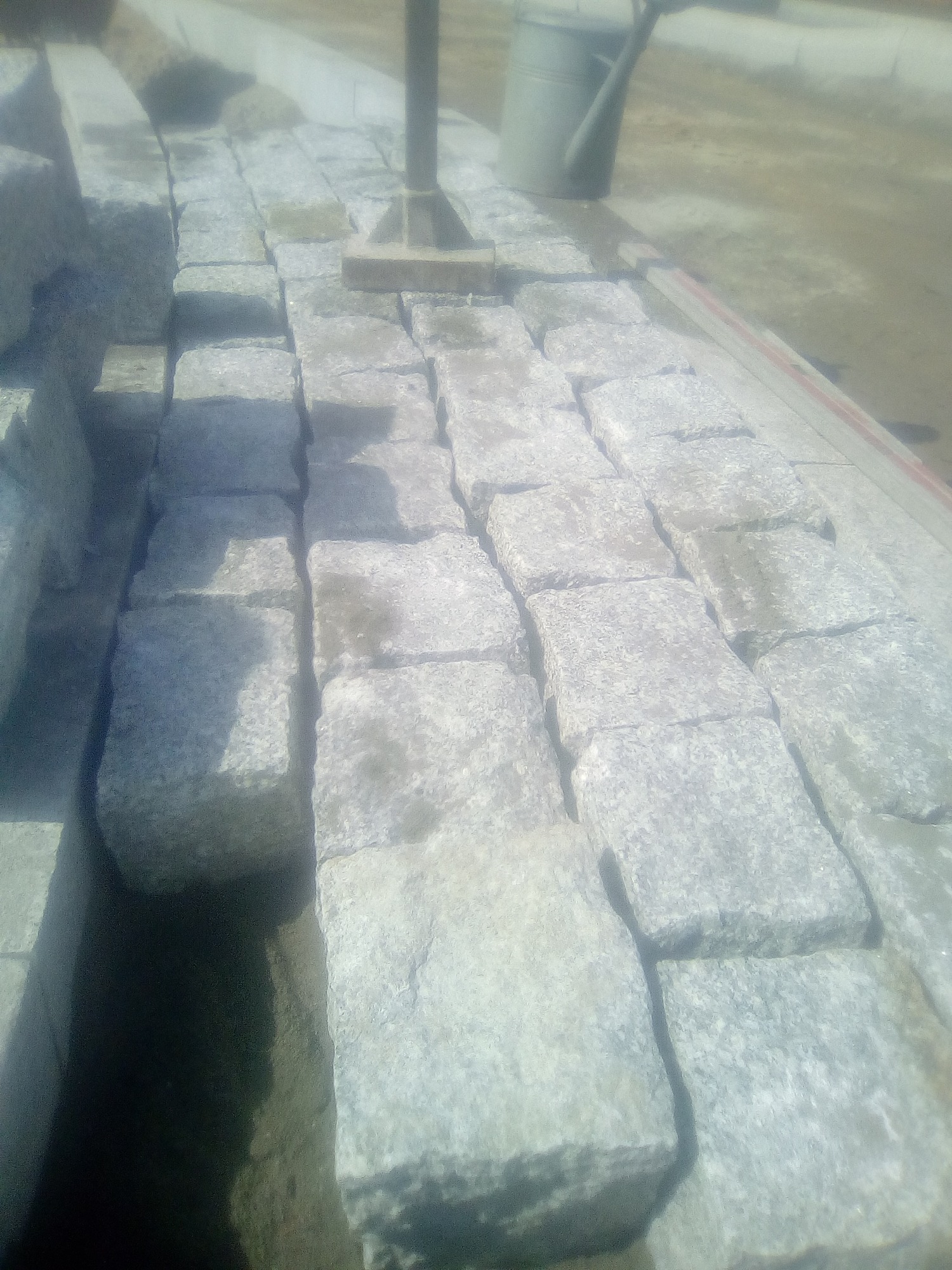 Szare kostki granitowe ułożone w rzędach, widoczna poziomica i konewka w tle, prawdopodobnie w trakcie prac brukarskich.