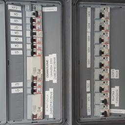 El-Ad - Otwarta skrzynka elektryczna z widocznymi bezpiecznikami i wyłącznikami, oznaczona etykietami opisującymi obwody: klimatyzacja, oświetlenie, gniazda 230V, złącze kablowe, szafa rack, klatka...