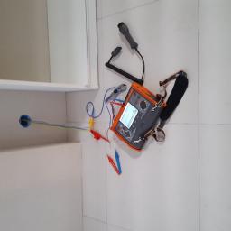 El-Ad - Miernik elektryczny z przewodami podłączonymi do gniazdka w ścianie, obok szafki w Krasnystawie, na białych płytkach podłogowych.