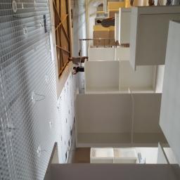 El-Ad - Montaż białych, modułowych regałów na tle ściany z metalową siatką i widocznymi pracownikami na drabinie.