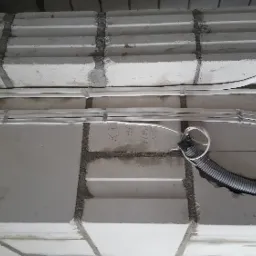 Białe przewody elektryczne przymocowane do surowego, szarego sufitu z bloczków betonowych za pomocą metalowych uchwytów, widoczne odręczne notatki na ścianie.