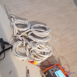 El-Ad - Zestaw białych przewodów zwiniętych w kręgi leżący na podłodze z płytek, obok torba narzędziowa i miernik elektryczny z przewodami zakończonymi krokodylkami. W tle widoczne nogi osoby...