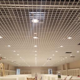 El-Ad - Montaż sufitu podwieszanego z oświetleniem punktowym w dużym pomieszczeniu; pracownik na drabinie reguluje lampę, widoczne otwarte przestrzenie w kratownicy sufitowej.