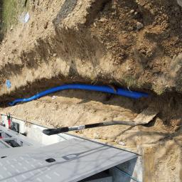 El-Ad - Niebieska rura karbowana ułożona w wykopie ziemnym, obok leży łopata z czarną rączką, widoczny fragment budynku z białymi oknami i klimatyzatorem na ścianie.