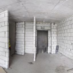 El-Ad - Wnętrze surowego mieszkania w stanie deweloperskim z widocznymi instalacjami elektrycznymi prowadzonymi po ścianach z betonu komórkowego, okna z widokiem na miasto i kable w peszlach leżące przy...