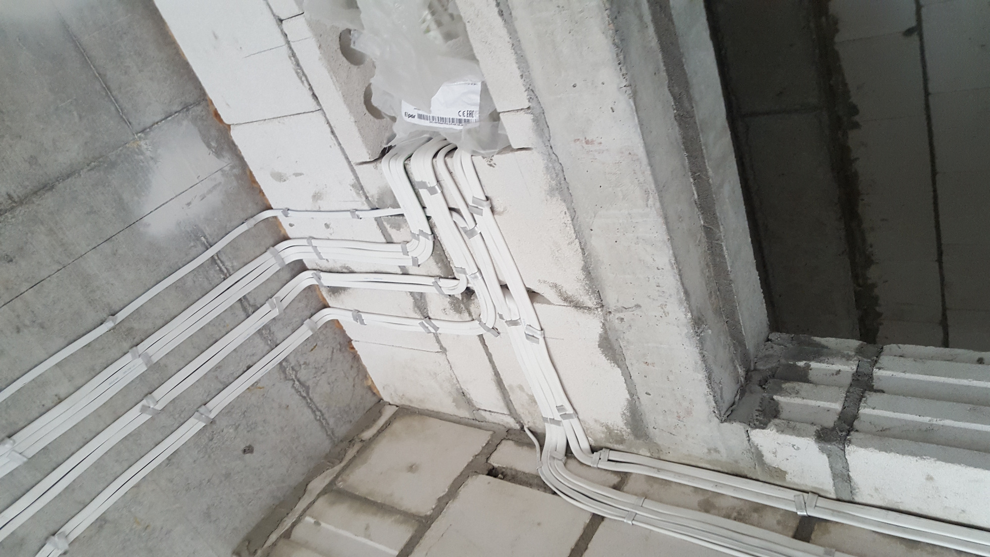 Białe kable elektryczne ułożone wzdłuż surowego, betonowego sufitu i ścian w trakcie instalacji, zabezpieczone szarą taśmą.