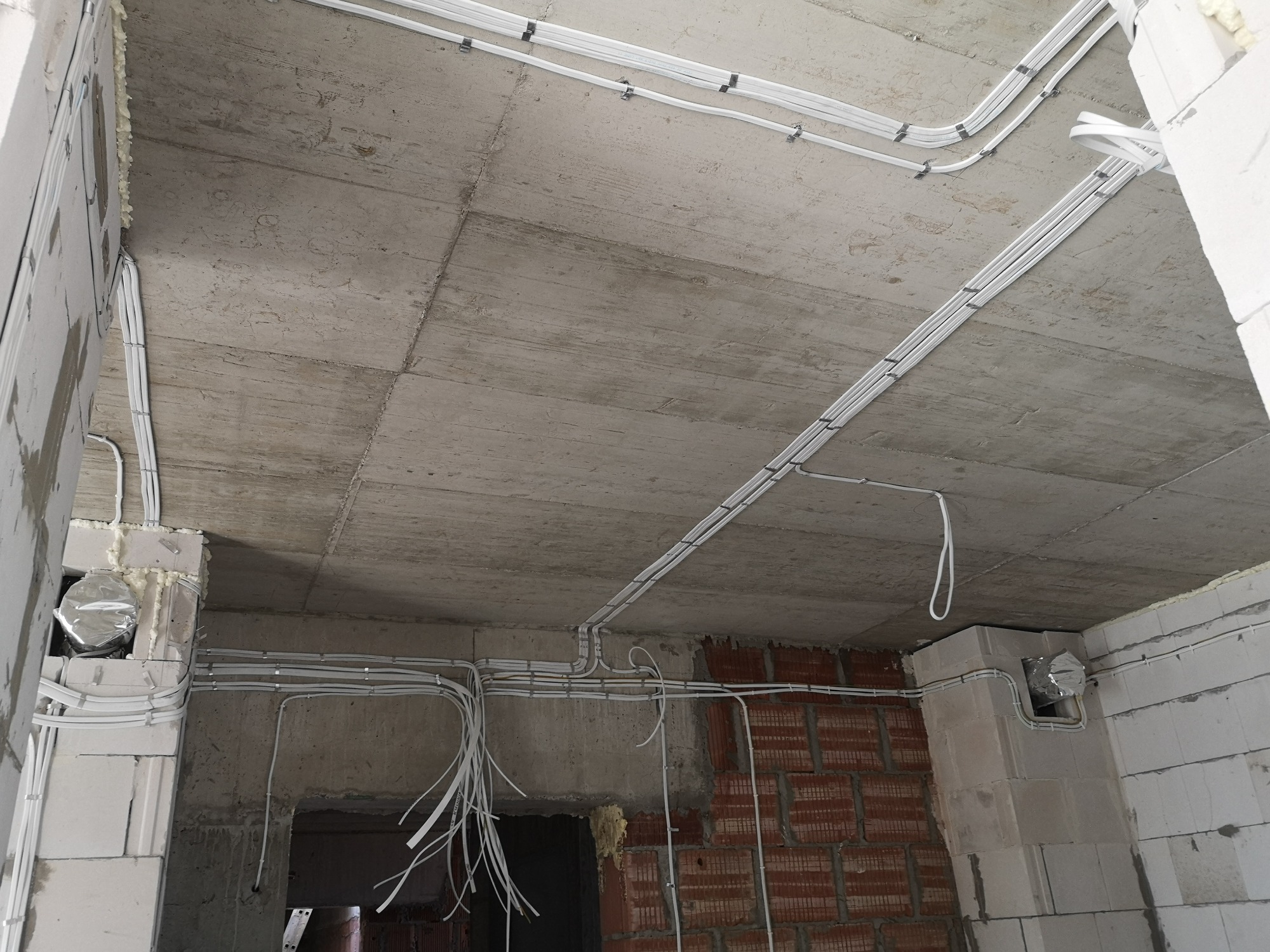 Białe kable elektryczne prowadzone po betonowym suficie i ścianach w surowym wnętrzu budynku, widoczne mocowania i luźno zwisające końcówki kabli.