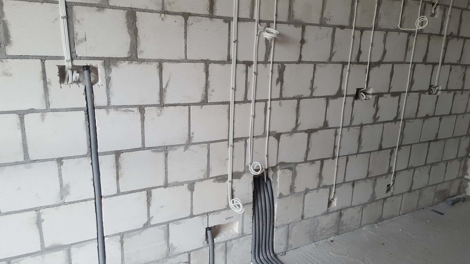 Surowa ściana z betonu komórkowego z widocznymi instalacjami elektrycznymi w białych i szarych peszlach, częściowo zamontowanymi w ścianie, z widocznymi puszkami instalacyjnymi.