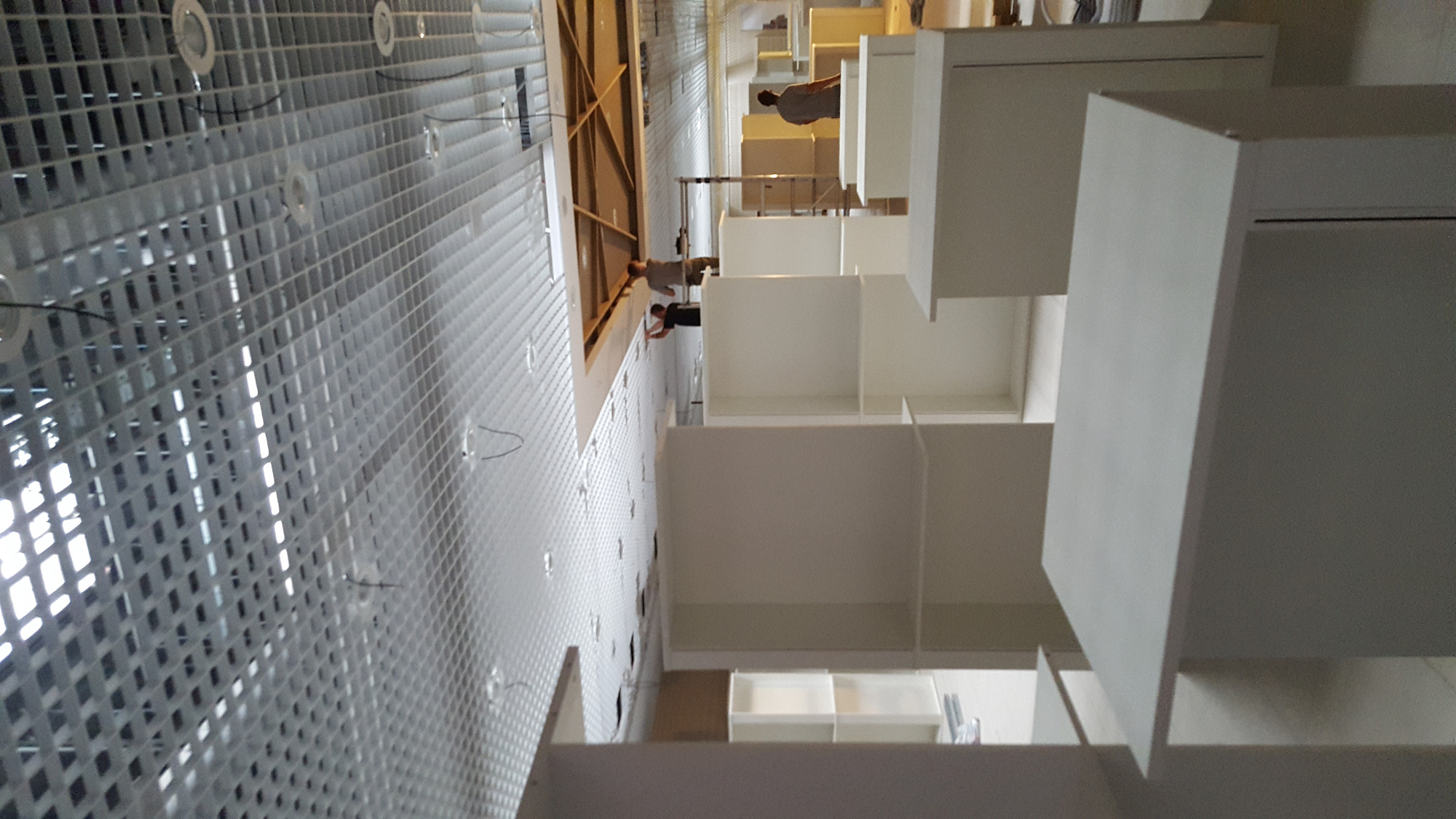 Montaż białych, modułowych regałów na tle ściany z metalową siatką i widocznymi pracownikami na drabinie.