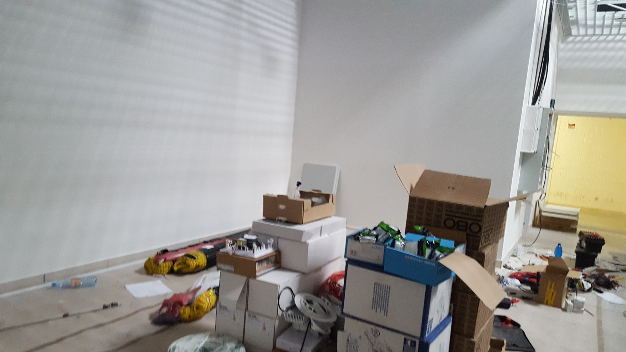 Wnętrze w trakcie prac instalacyjnych, widoczne kartony z osprzętem elektrycznym OBO, rozłożone kable, narzędzia i puszki instalacyjne na podłodze, surowe ściany i żółte pomieszczenie w tle.