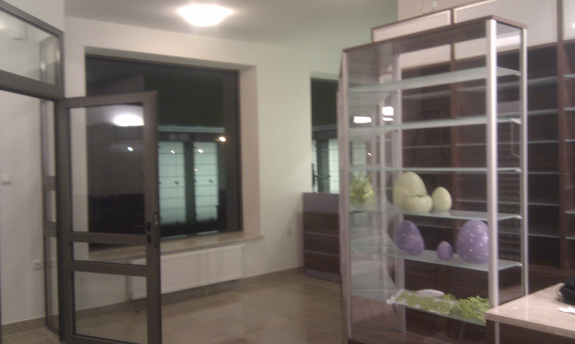 Wnętrze z otwartymi szklanymi drzwiami, oknem z widokiem na noc oraz szklaną witryną z dekoracjami w kształcie jajek i zielonymi roślinami.