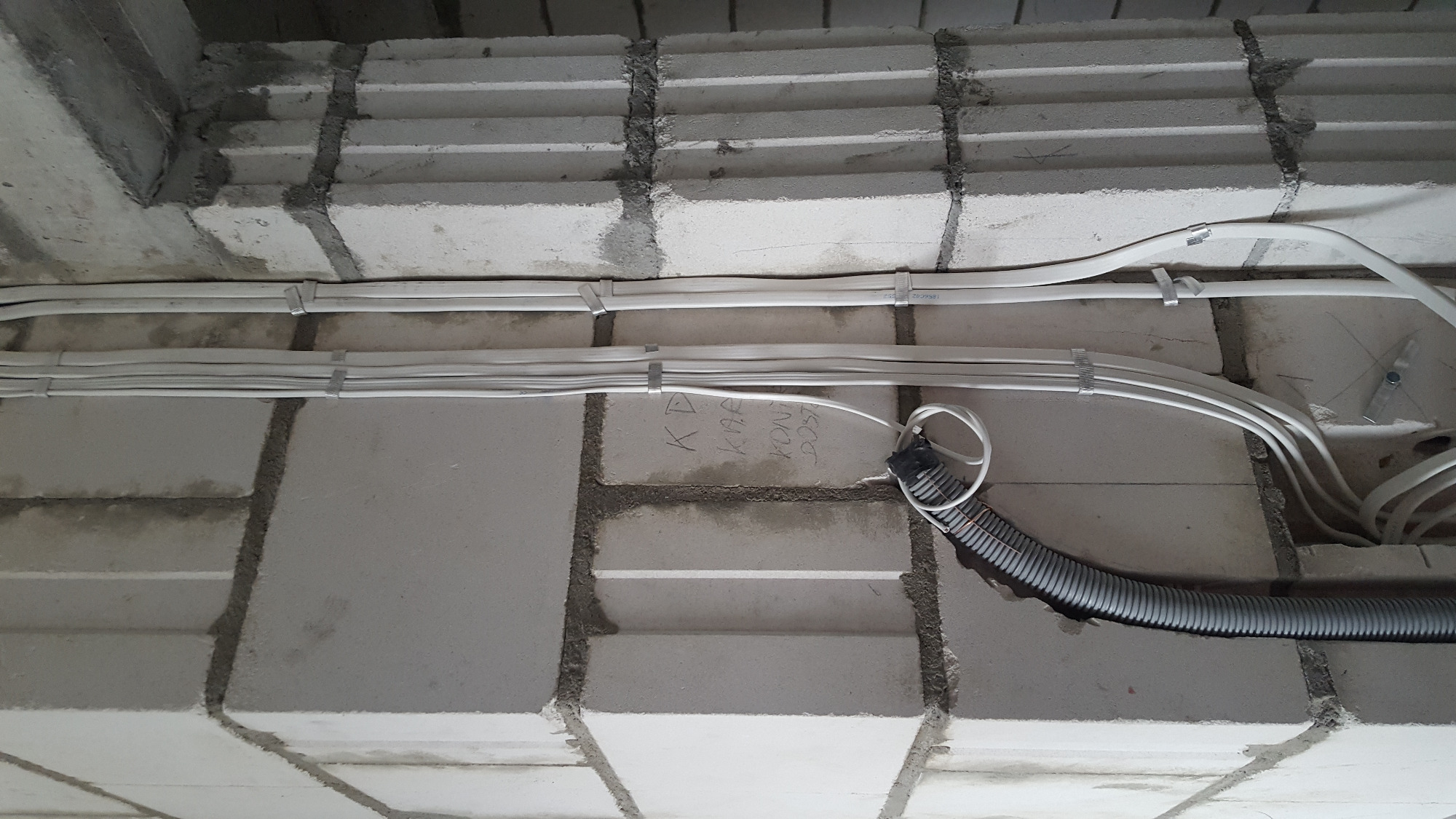 Białe przewody elektryczne przymocowane do surowego, szarego sufitu z bloczków betonowych za pomocą metalowych uchwytów, widoczne odręczne notatki na ścianie.