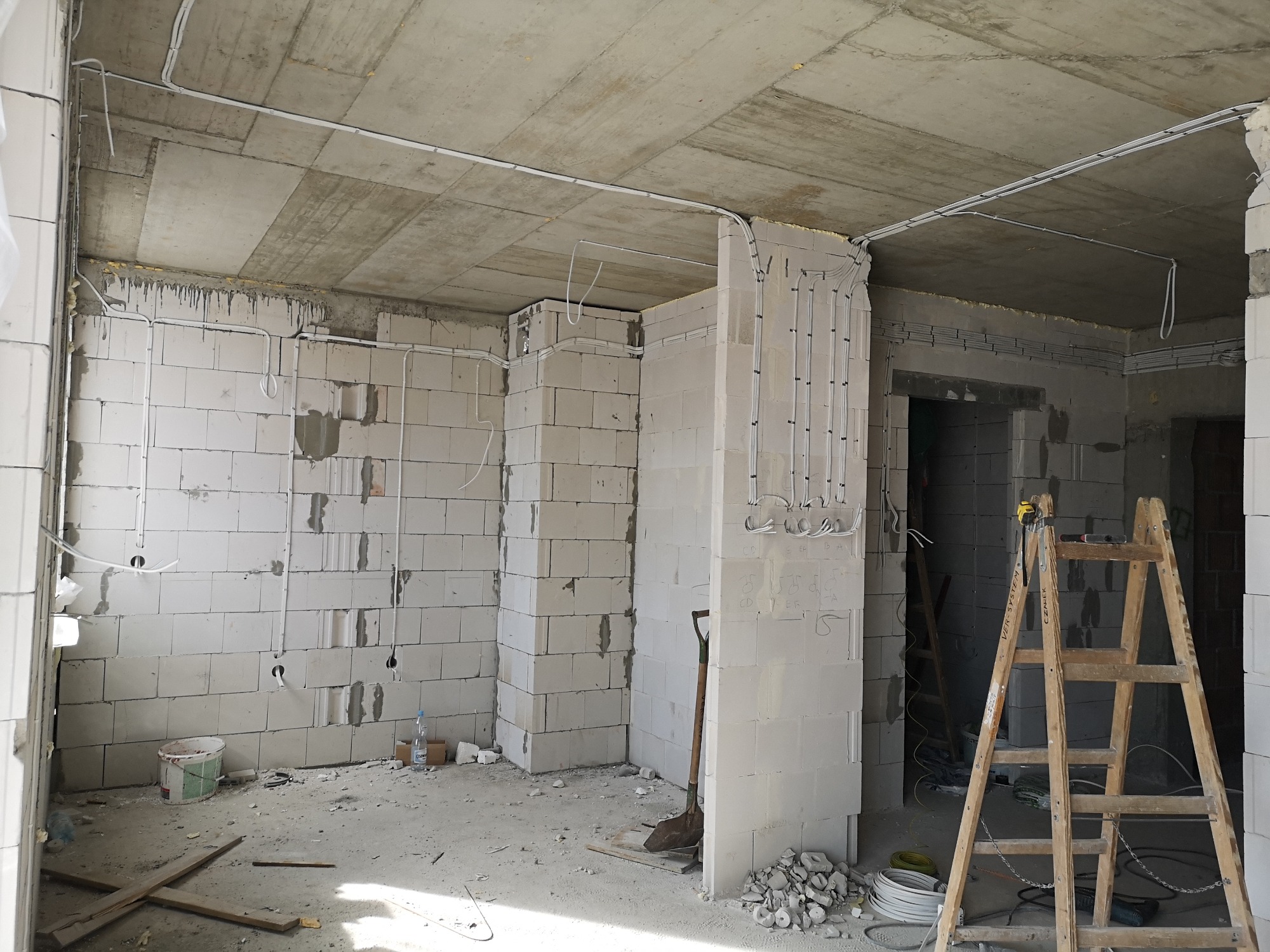 Surowy stan wnętrza z instalacją elektryczną w białych rurkach, ściany z białych bloczków, drewniana drabina oparta o ścianę, widoczne przewody elektryczne na suficie i ścianach, w tle przejście...